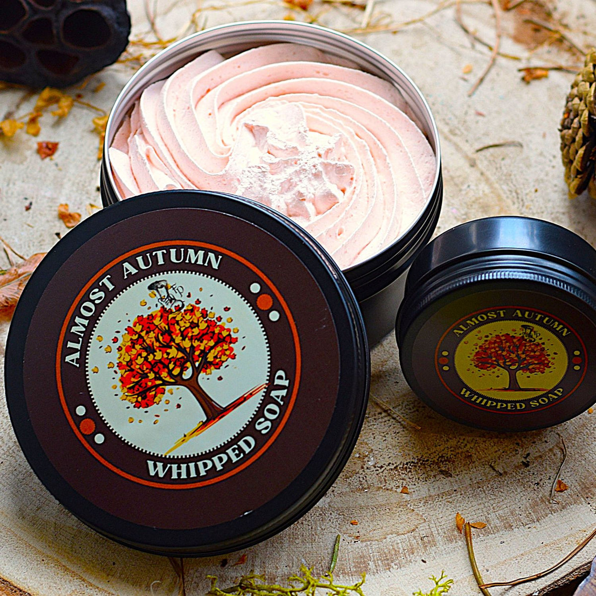 A tin of whipped soap shown open with pale orange whipped soap inside, alongside two closed tins. The lids feature a circular autumn tree label reading 'ALMOST AUTUMN WHIPPED SOAP.