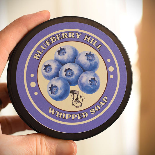 A hand holding a round tin of Blueberry Hill Whipped Soap. The tin features a purple and beige label with an illustration of blueberries in the center.