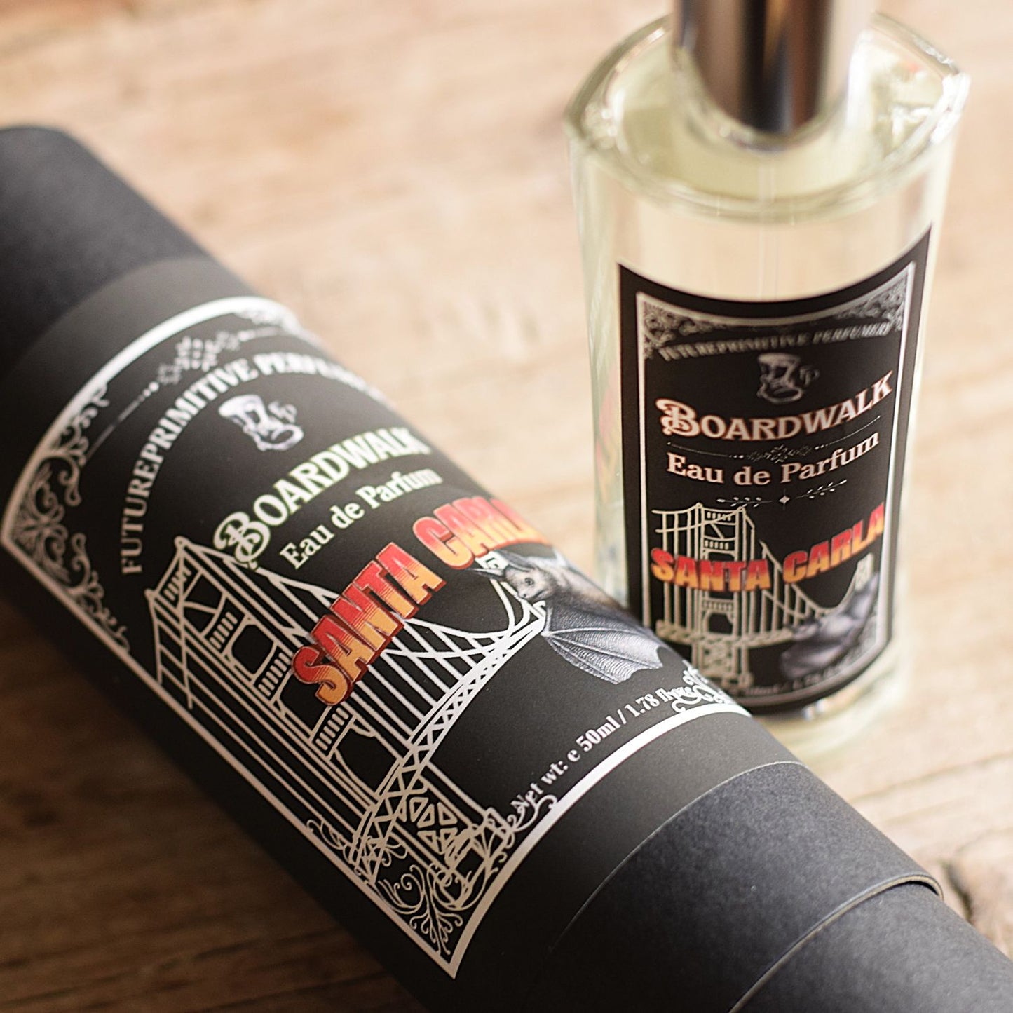 A bottle of Boardwalk Eau de Parfum next to its black cylindrical packaging tube, both featuring white and red graphics, set on a light wooden surface.
