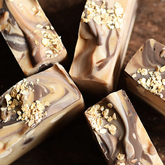 Several rectangular bars of marbled chocolate and oat cookie butter soap with crushed oats on top, arranged on a dark wooden surface.