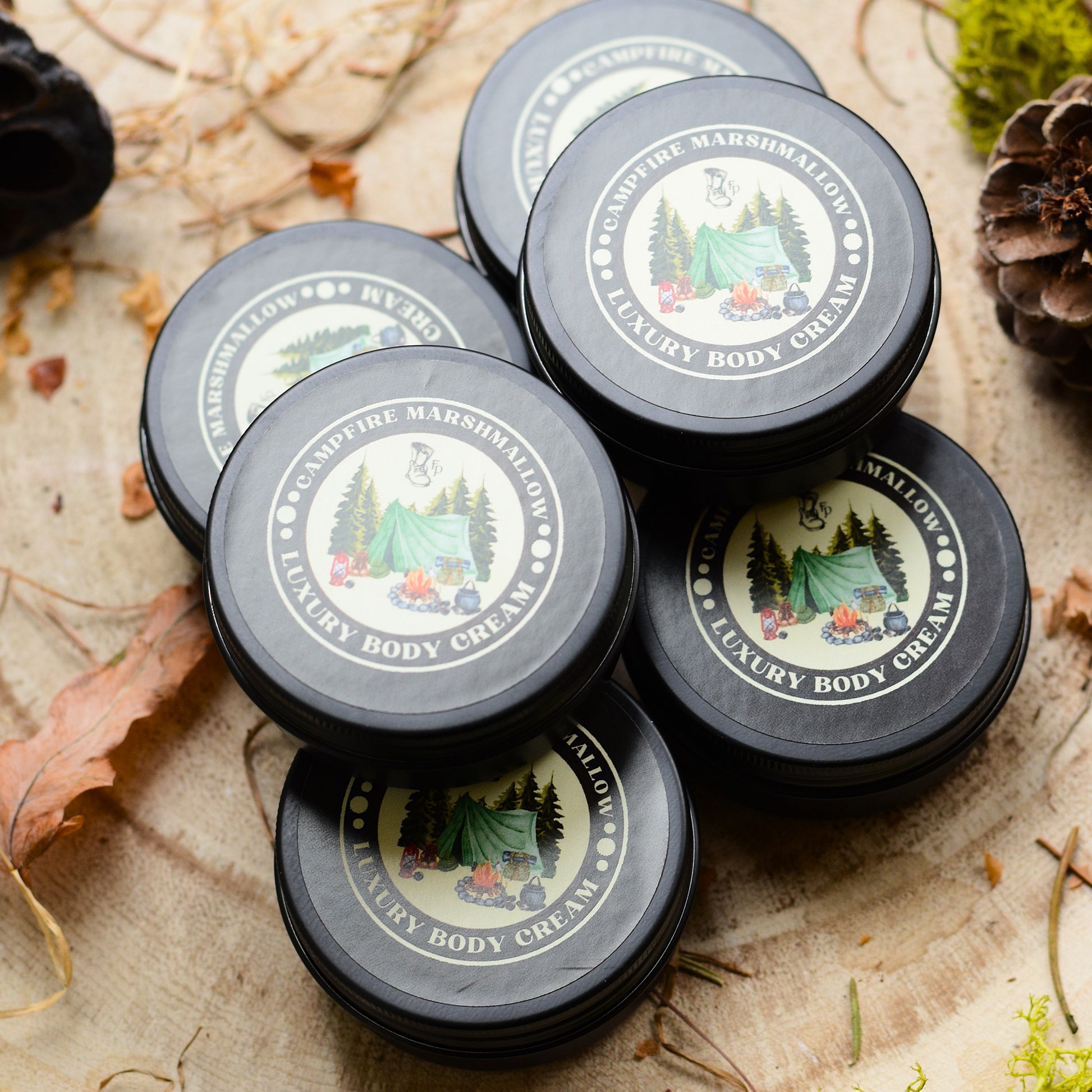 Multiple jars of 'Campfire Marshmallow' luxury body cream on a natural background with leaves and pine cones.