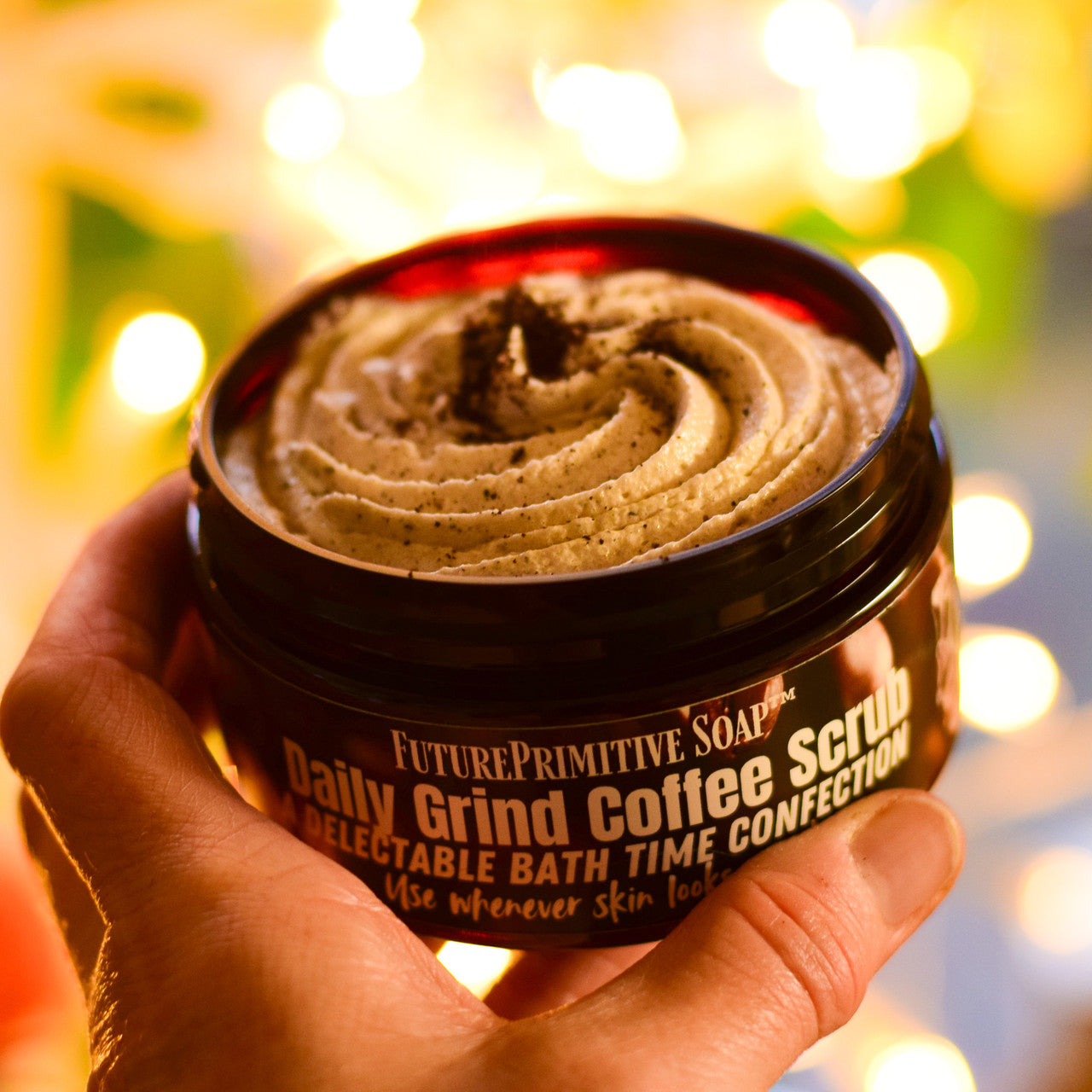 Jar of Future Primitive Soap Daily Grind Coffee Scrub held by a hand with a blurred lights background