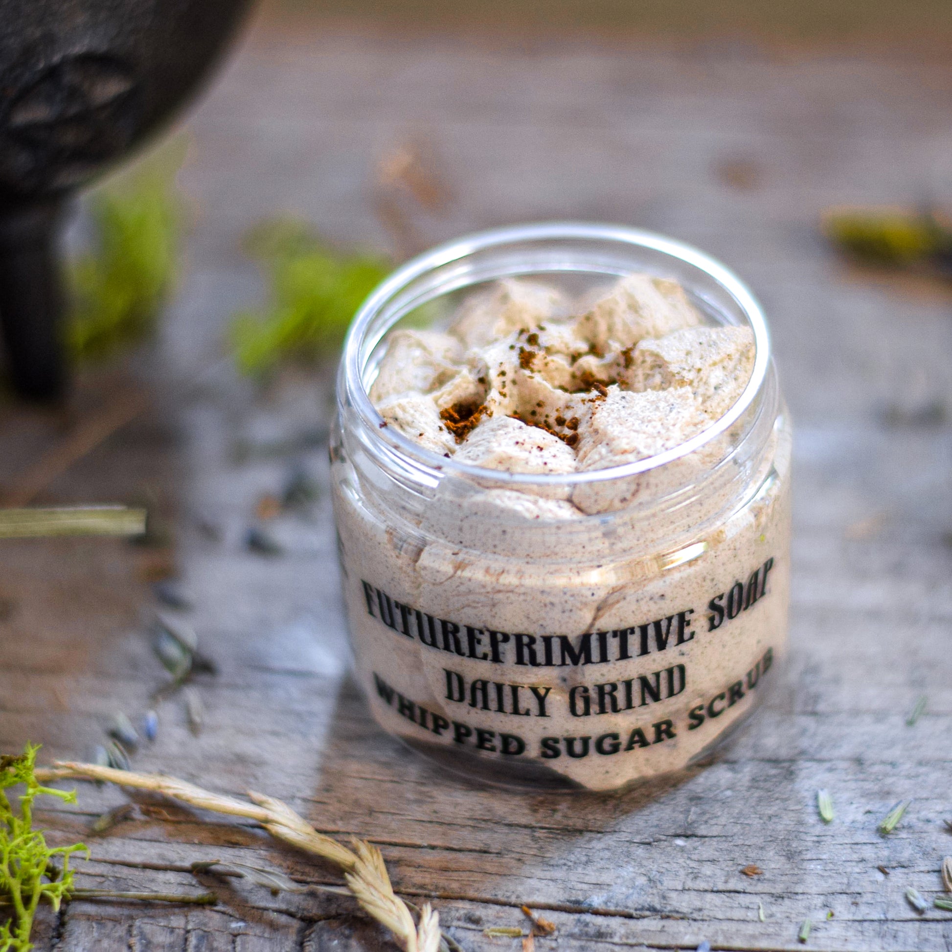 Jar of whipped sugar scrub labeled 'Futurereprimitive Soap Daily Grind' on a wooden surface with natural elements.