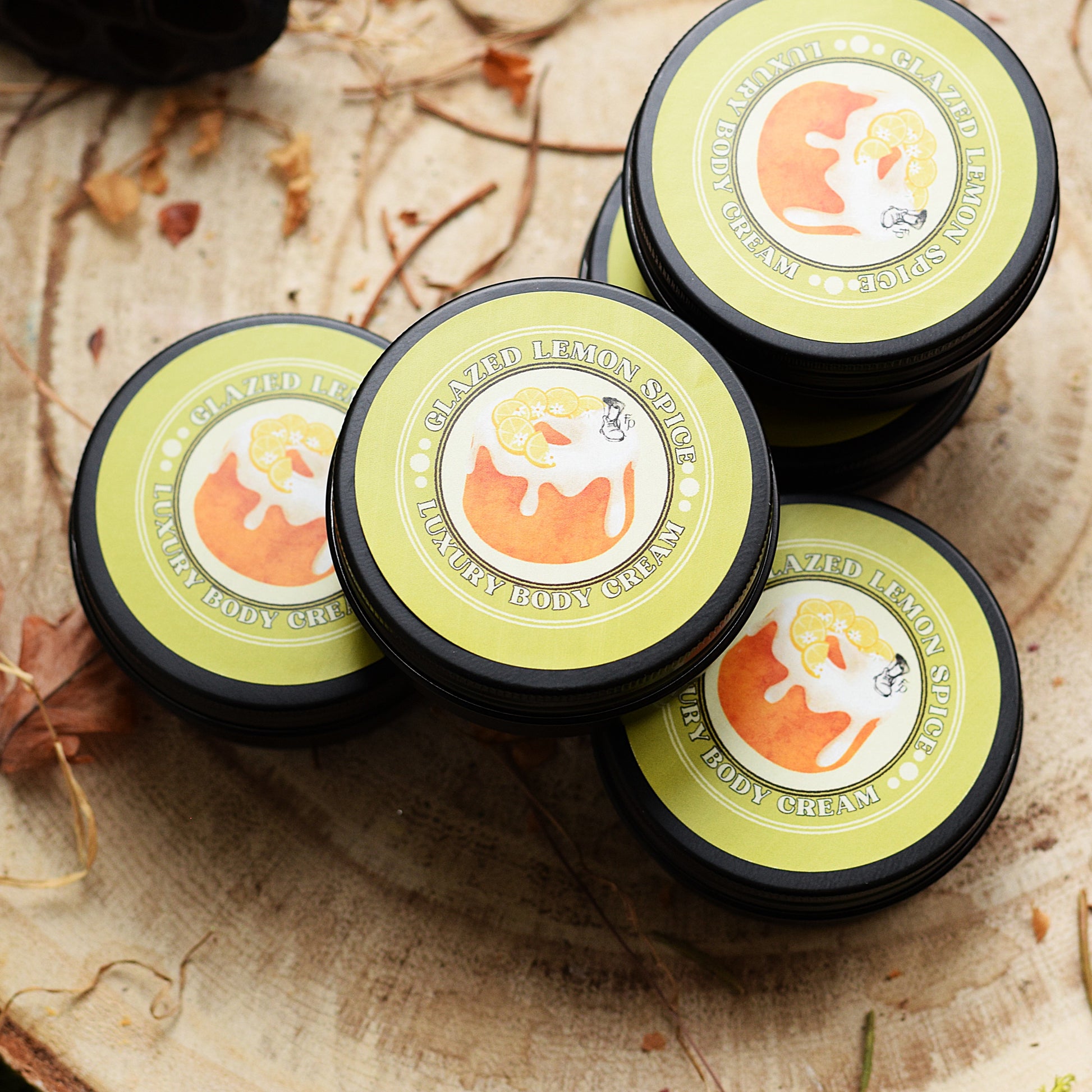 Four containers of lemon spice body cream on a wooden surface with leaves.