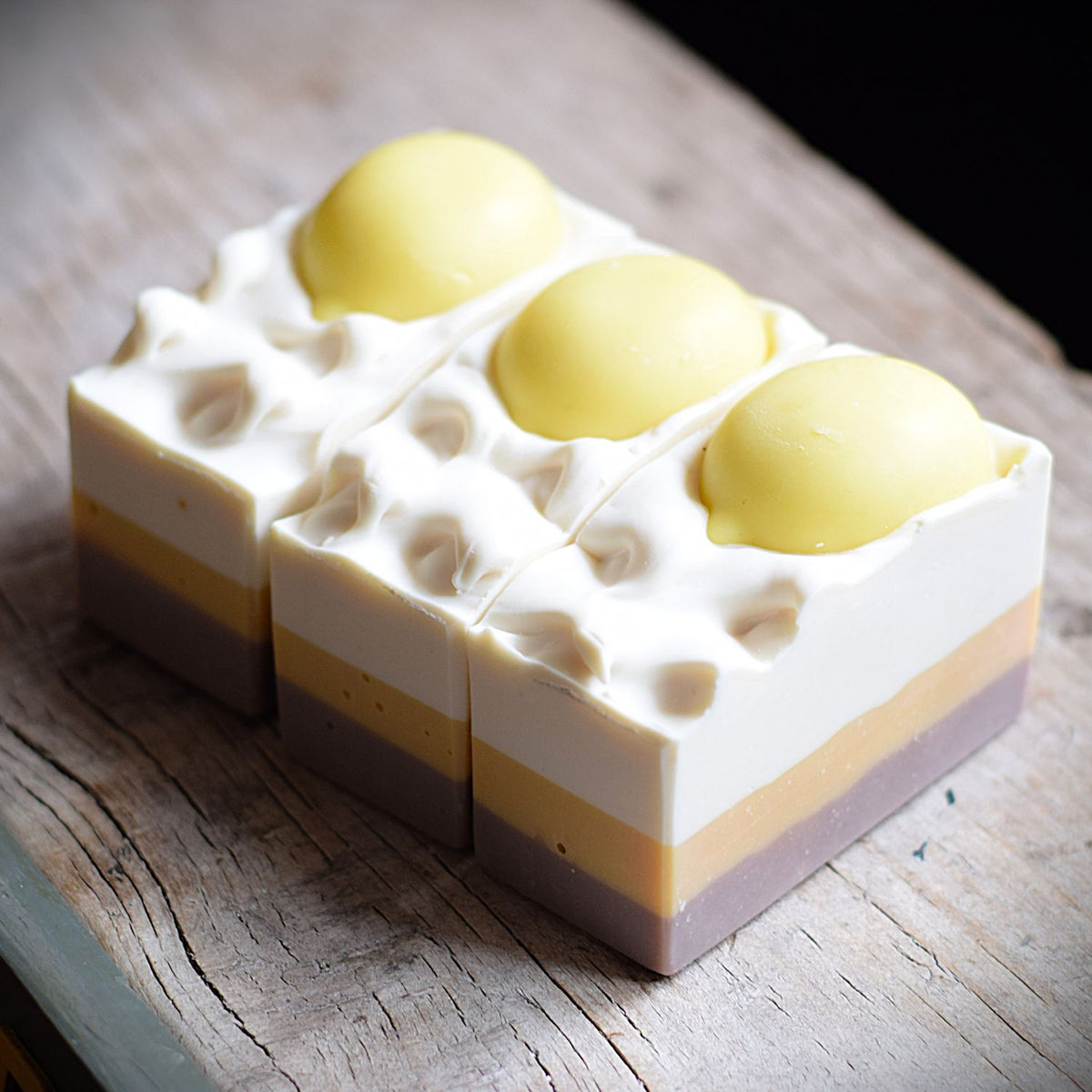 three handmade soaps with lemon soaps stuck into the topping. the soaps are sitting on a rustic wooden table.