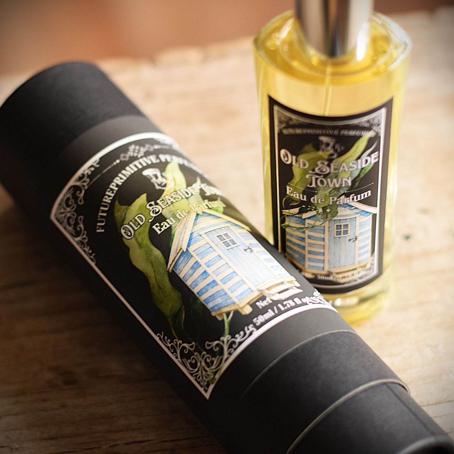 A bottle of Old Seaside Town Eau de Parfum standing next to its black cylindrical packaging tube on a rustic wooden surface. The label features a vintage illustration of a blue seaside town house and green foliage.