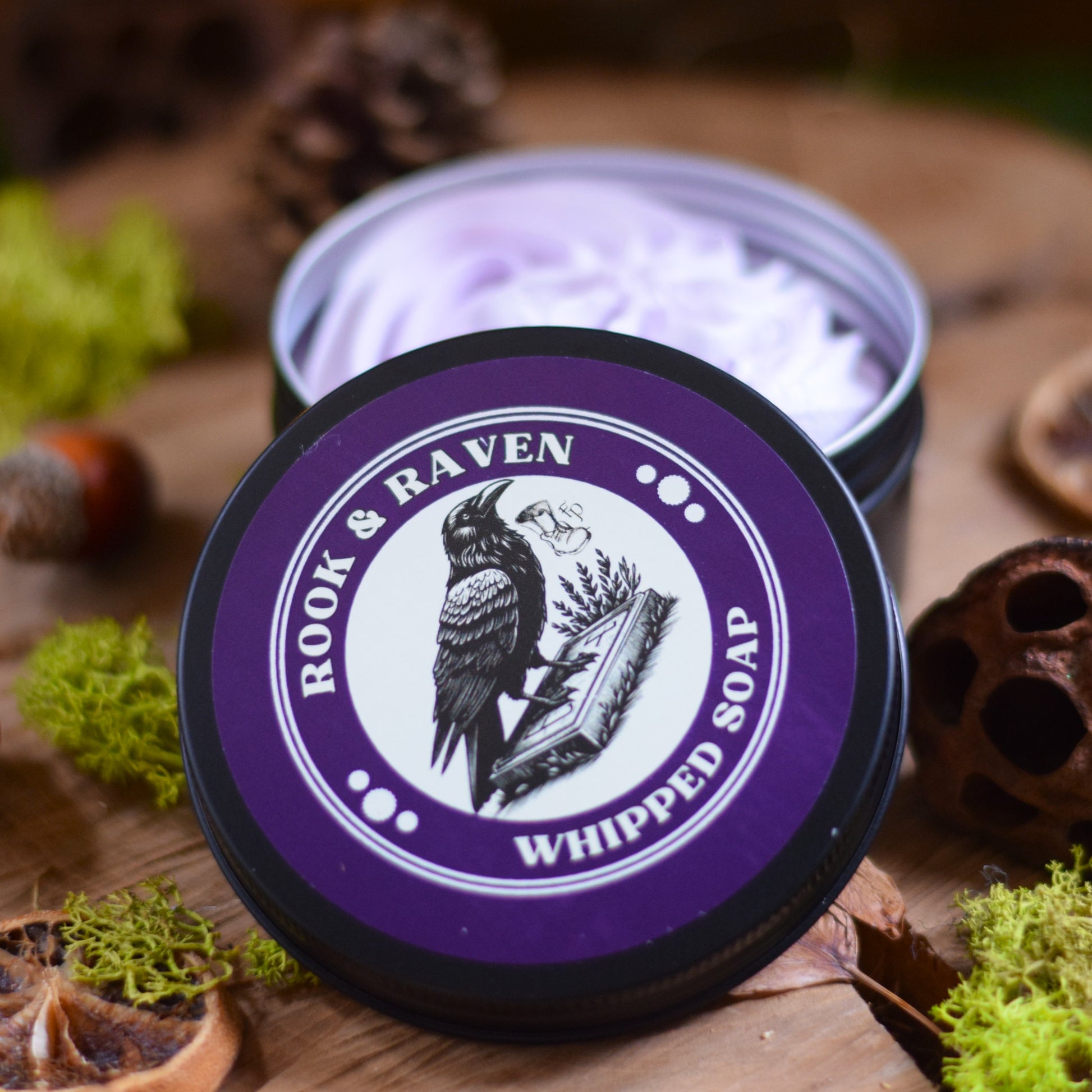 Container of Rook & Raven whipped soap on a wooden surface with natural elements.