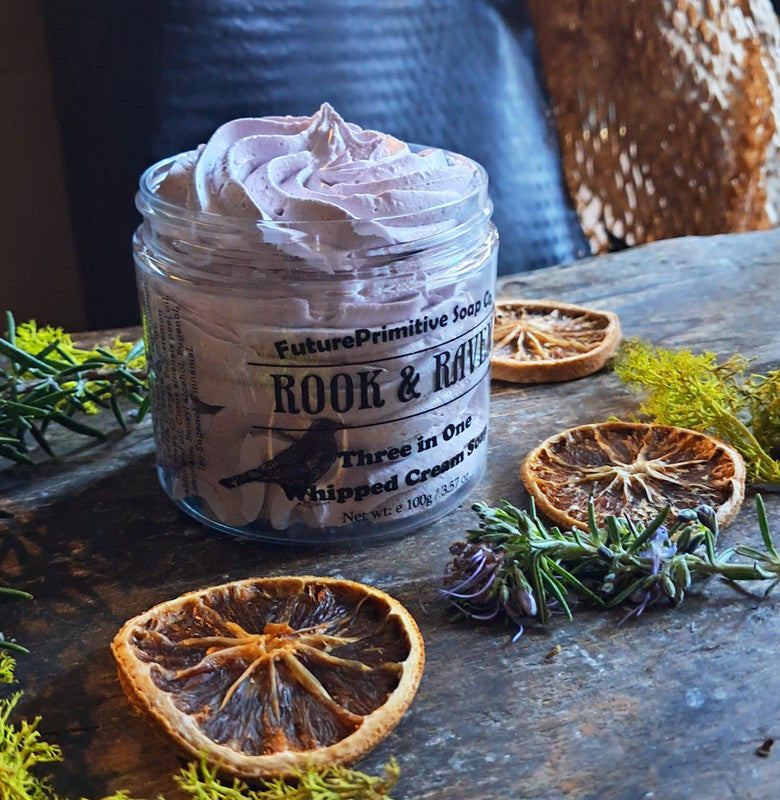 Jar of 'Rook & Raven' whipped cream soap on a rustic surface with dried oranges and herbs.