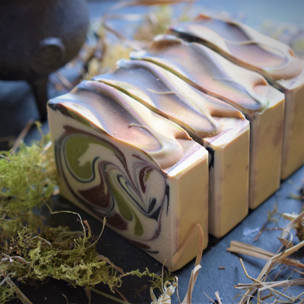 Four rectangular bars of artisanal soap with marbled swirls of cream, brown, purple, and olive green, arranged on a dark slate surface with dried grass and moss props.