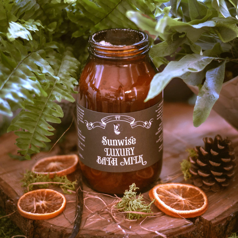 Brown glass jar labeled 'Sunwise Luxury Body Milk' on a wooden surface with greenery and oranges.
