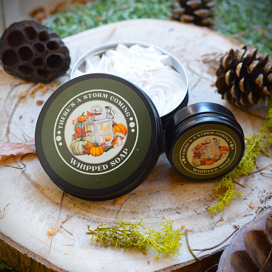 Two containers of 'There's a Storm Coming' whipped soap on a wooden surface with natural elements.