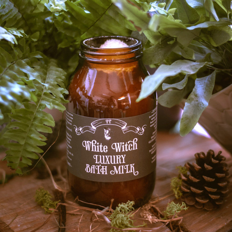 Brown bottle labeled 'White Witch Luxury Bath Milk' surrounded by greenery on a wooden surface