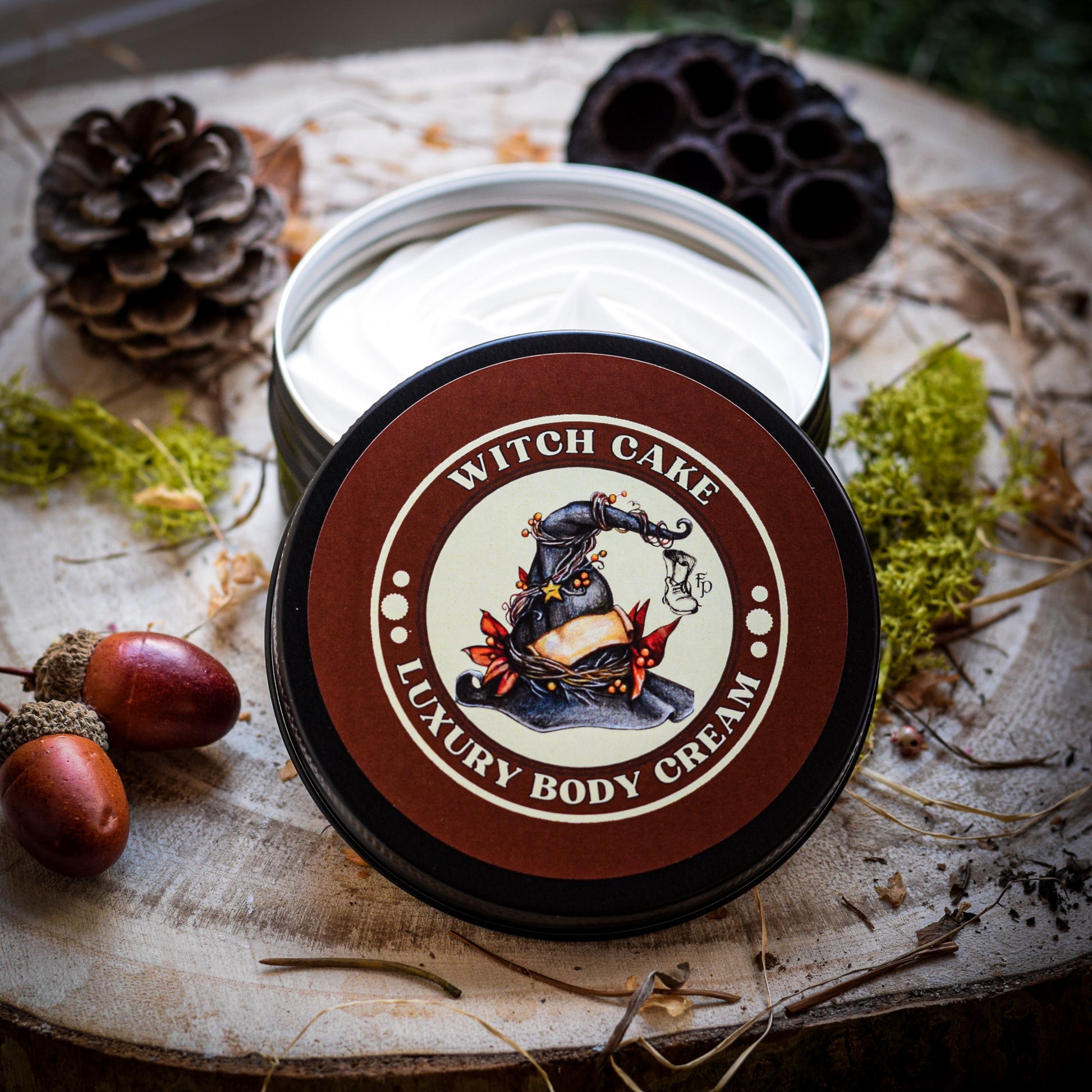 Witch Cake Luxury Body Cream sitting on a piece of wood surrounded by dried moss and seed pods