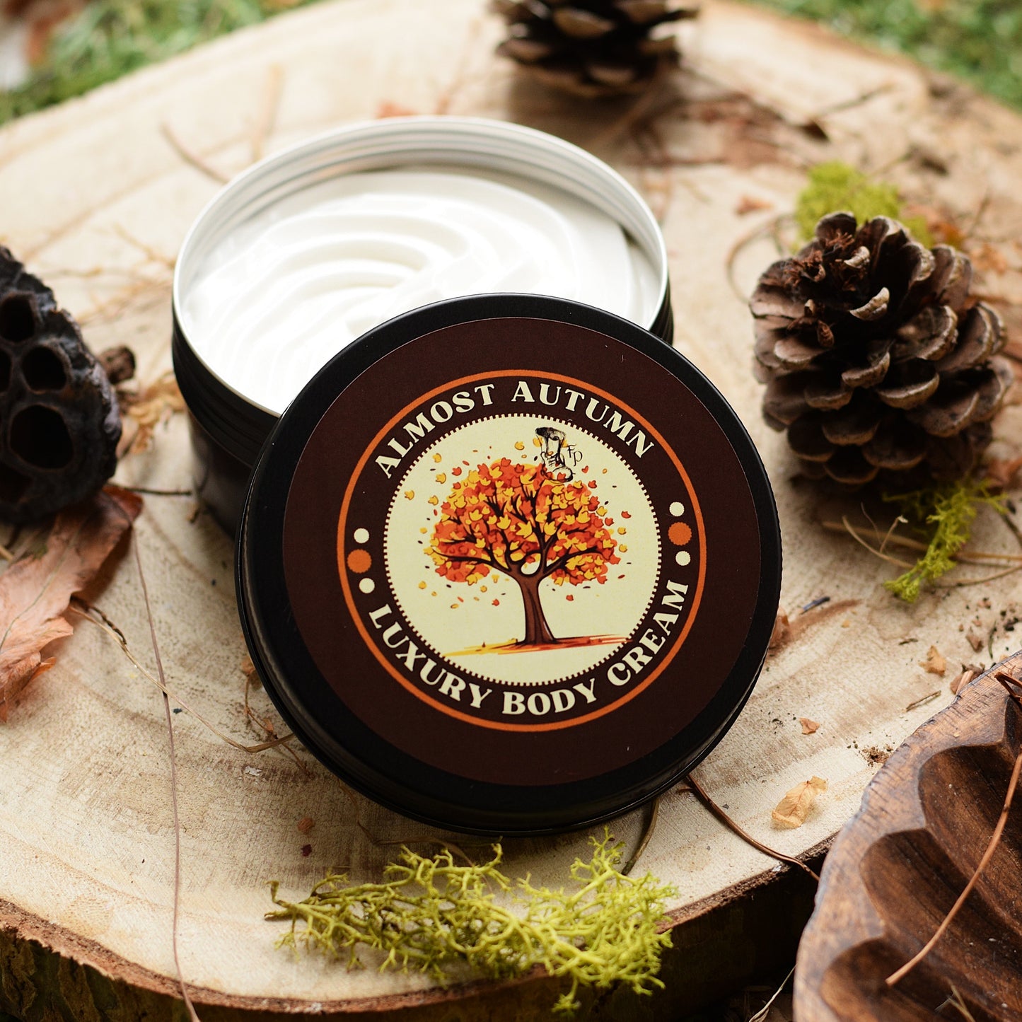 An open round tin of luxury body cream sits on a wood slice with pinecones and moss around it. The lid is placed in front and shows a label reading 'ALMOST AUTUMN LUXURY BODY CREAM' with an autumn tree illustration.