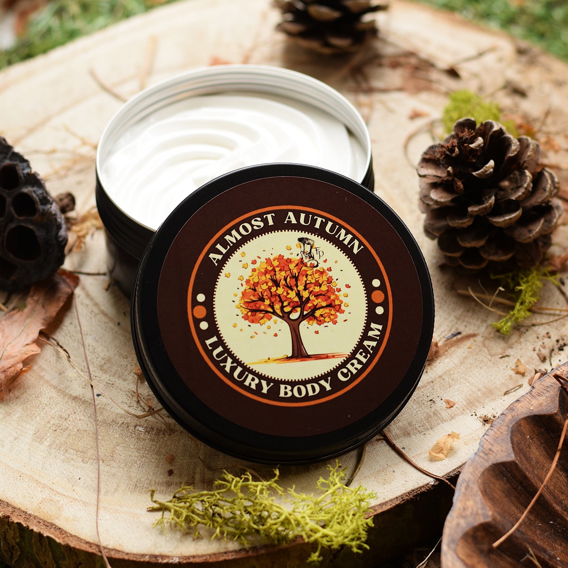 An open round tin of luxury body cream sits on a wood slice with pinecones and moss around it. The lid is placed in front and shows a label reading 'ALMOST AUTUMN LUXURY BODY CREAM' with an autumn tree illustration.