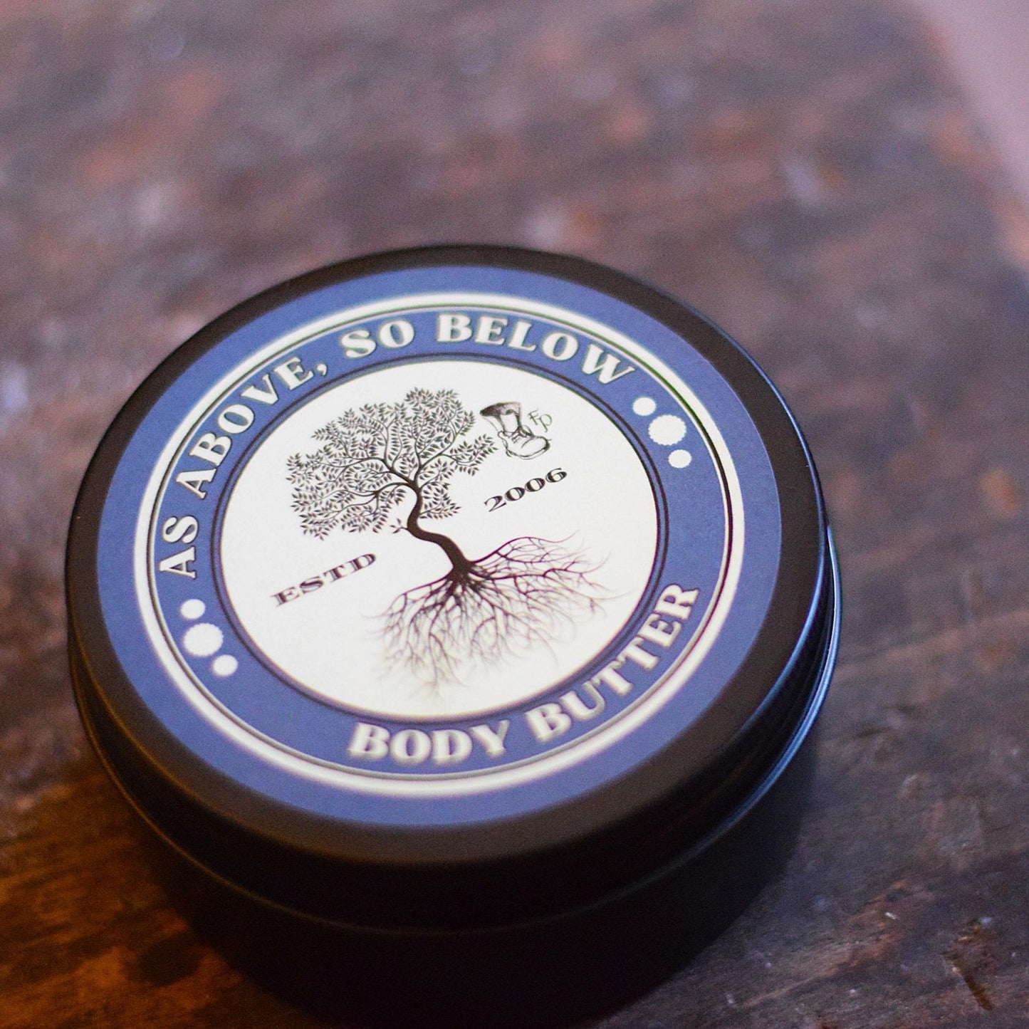 Body butter container with 'As Above, So Below' branding on a wooden surface