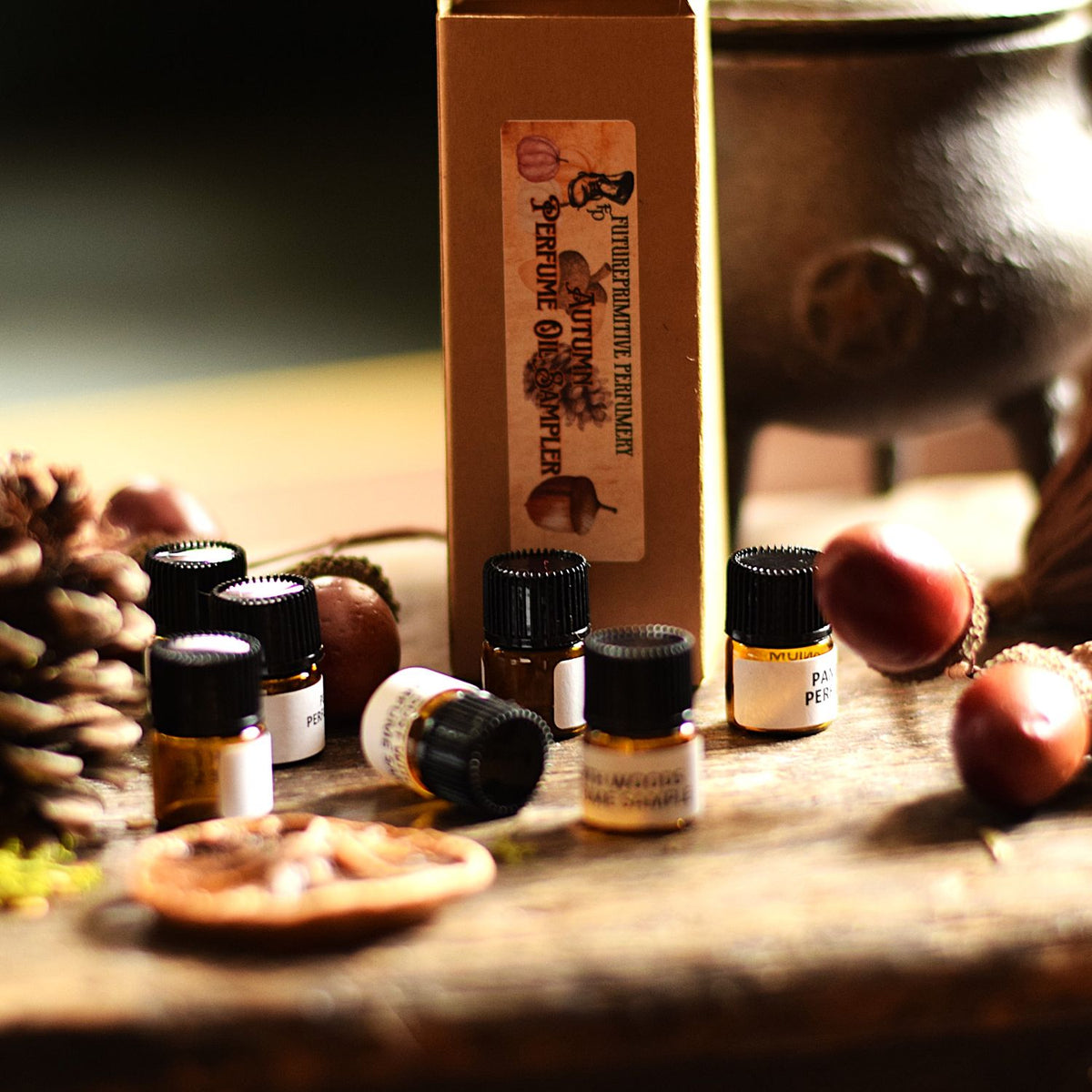 Essential oil bottles and a cardboard box labeled 'Autumn Perfume Oil Sampler' arranged on a rustic wooden surface with acorns, a pinecone, and a metal cauldron in the background.