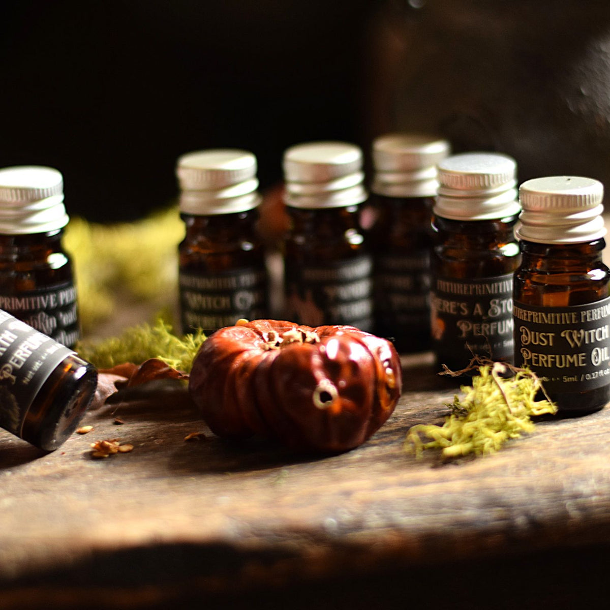 A group of amber glass essential oil bottles with silver caps and black labels, arranged on a rustic wooden surface with dried moss and a small red decorative object in the foreground.