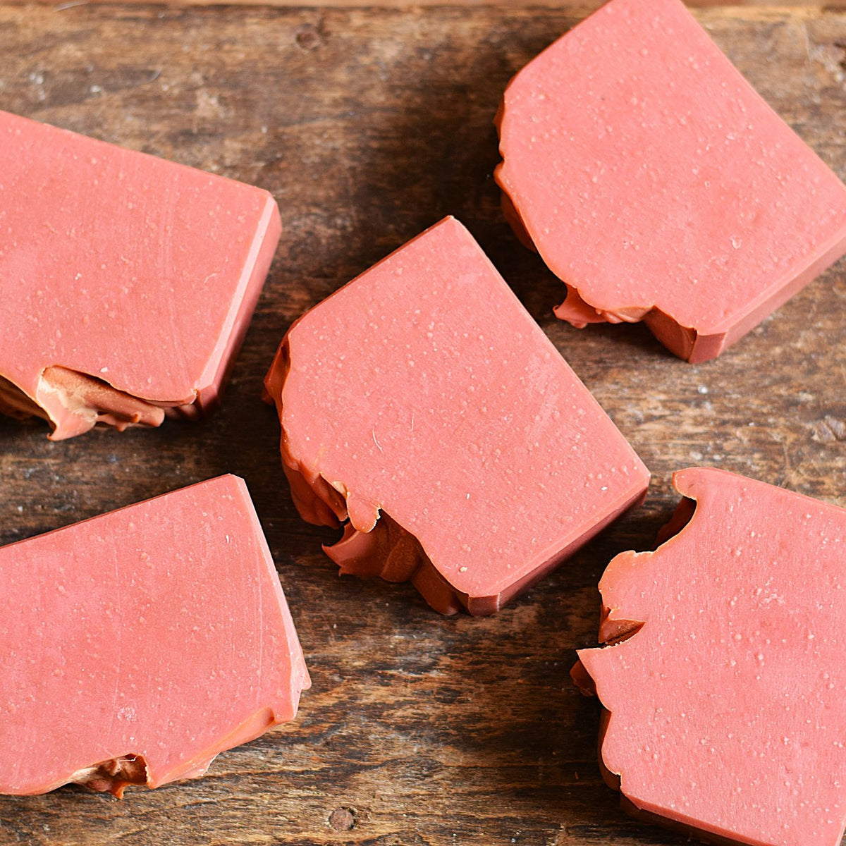Four rough-cut pinkish-red soap bars arranged on a rustic dark wooden surface.