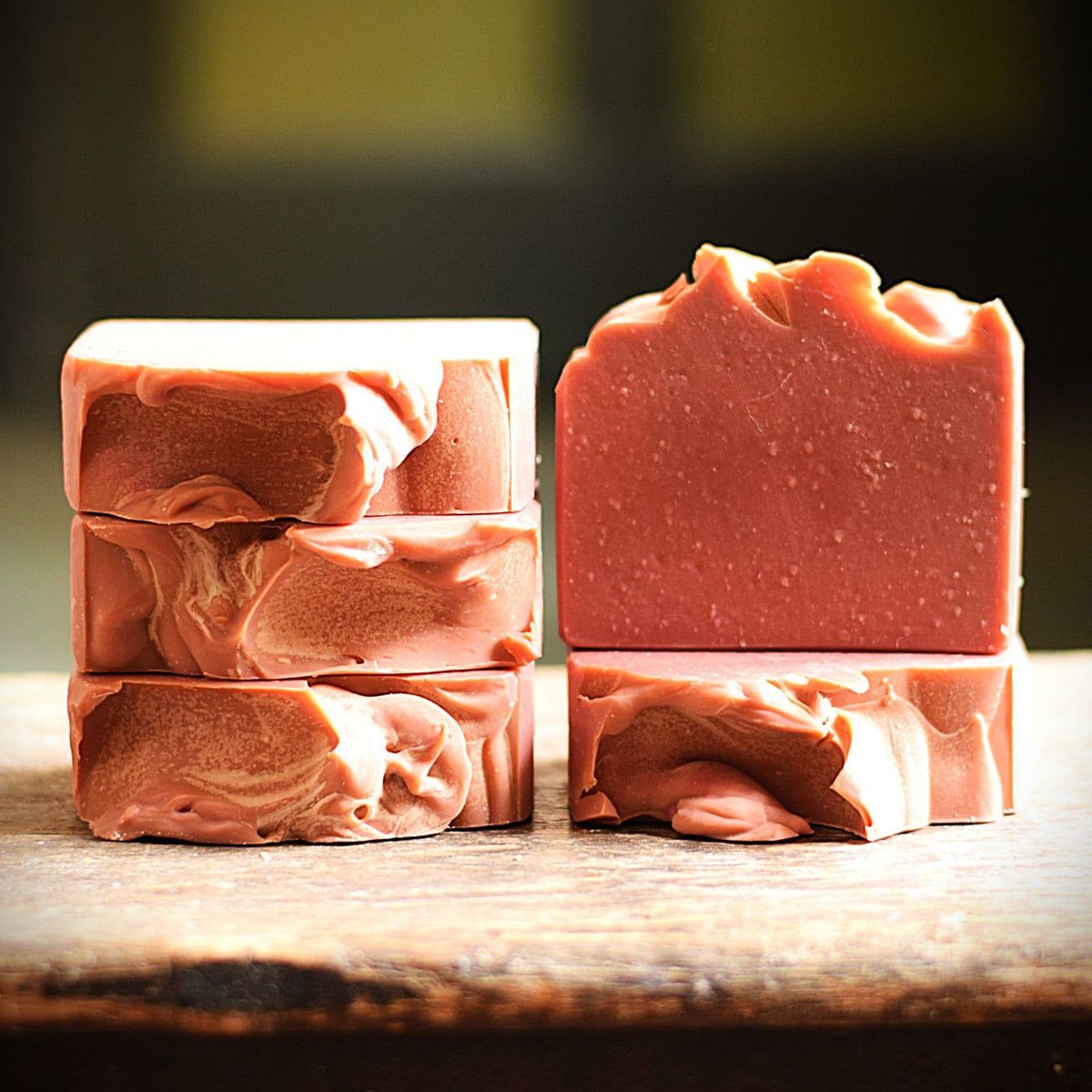 Three handmade soap bars with a dusty rose pink color, stacked on a rustic wooden surface with a dark, blurred background.
