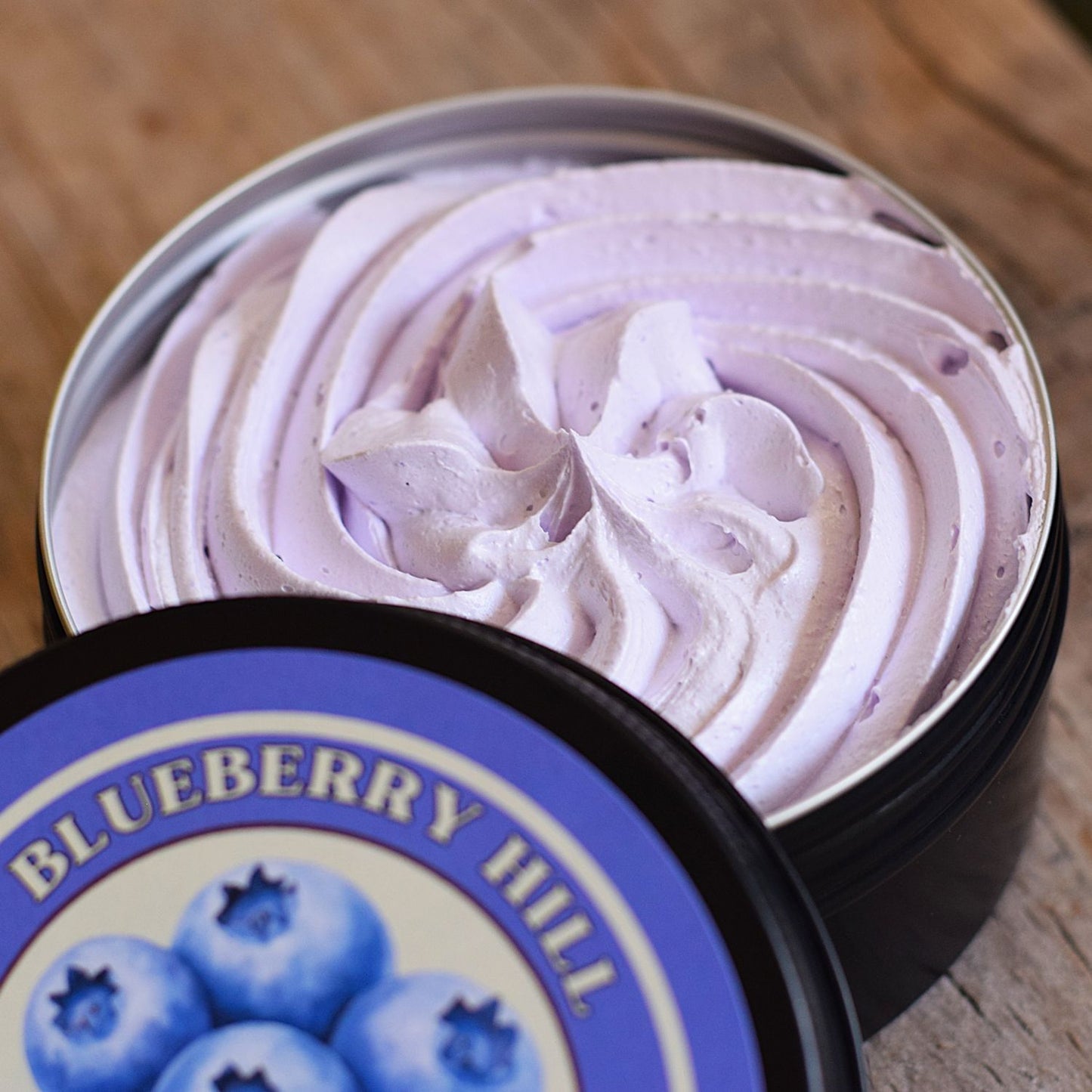 A close-up of a whipped lavender-pink cosmetic cream or balm swirled inside an open round metal tin, with the branded lid placed in front on a wooden surface. The label reads Blueberry Hill and shows blueberries.