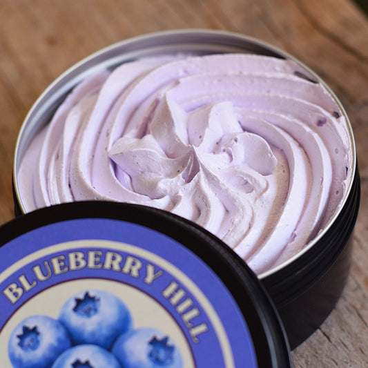 A close-up of a whipped lavender-pink cosmetic cream or balm swirled inside an open round metal tin, with the branded lid placed in front on a wooden surface. The label reads Blueberry Hill and shows blueberries.