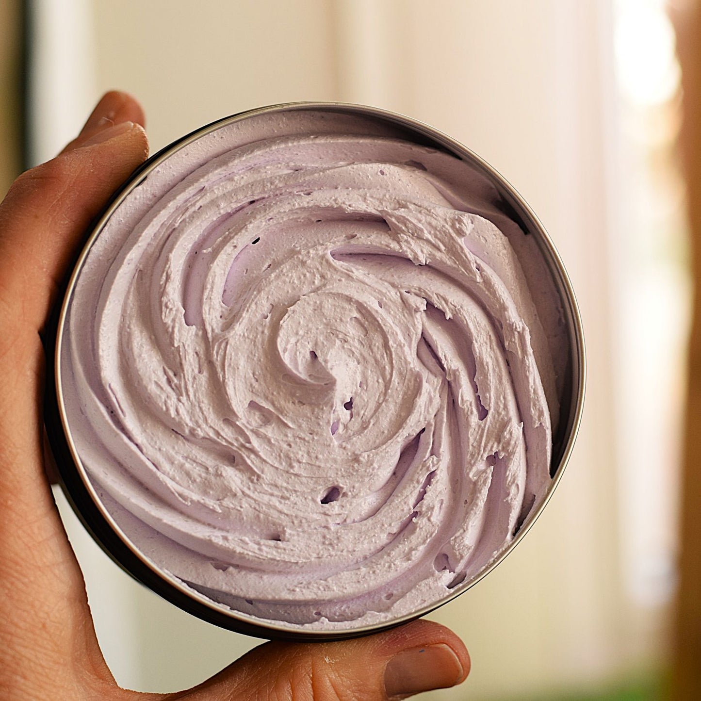 A close-up of a hand holding a round metal tin filled with whipped lavender-colored cream or balm, shown against a softly blurred indoor background.