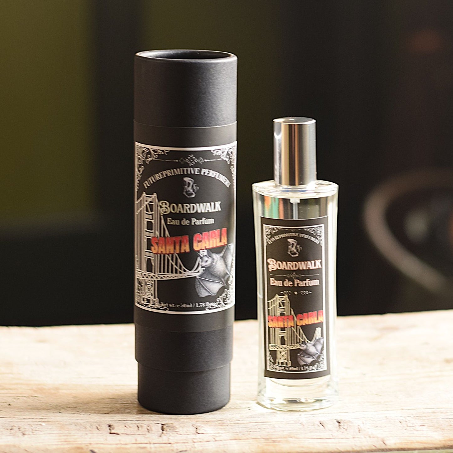 A bottle of Boardwalk Eau de Parfum next to its black cylindrical packaging tube, displayed on a light wooden surface with a dark, blurred background.