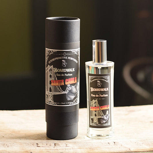 A bottle of Boardwalk Eau de Parfum next to its black cylindrical packaging tube, displayed on a light wooden surface with a dark, blurred background.