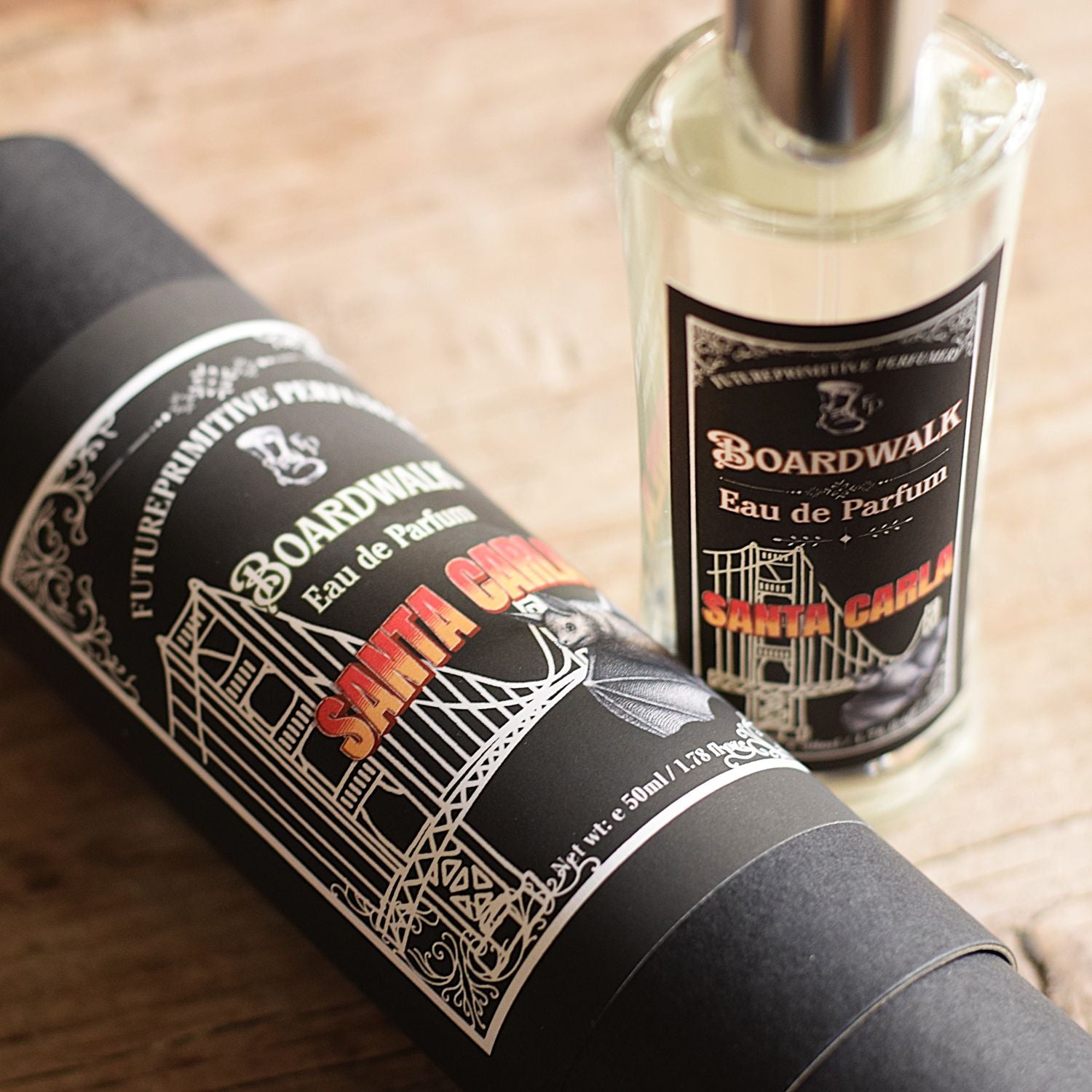 A bottle of Boardwalk Eau de Parfum next to its black cylindrical packaging tube, both featuring white and red graphics, set on a light wooden surface.