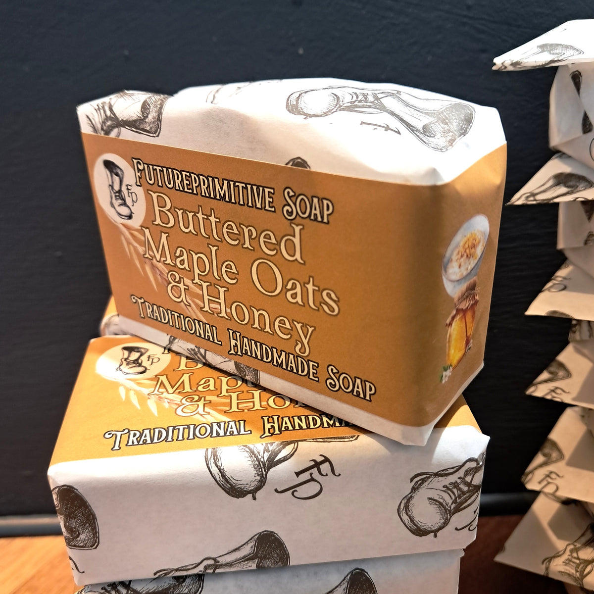 Two wrapped bars of Future Primitive Soap, Buttered Maple Oats & Honey, stacked on top of each other. The packaging features a warm tan and white color scheme with black illustrations of a bowl and spoon, set against a dark background.