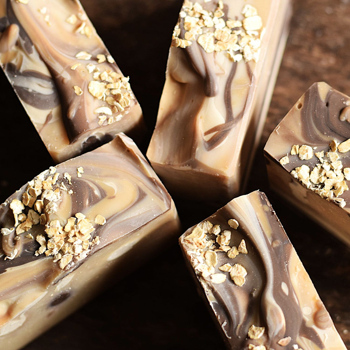 Several rectangular bars of marbled chocolate and oat cookie butter soap with crushed oats on top, arranged on a dark wooden surface.