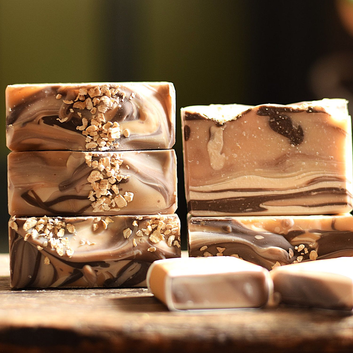 Several rectangular bars of marbled soap in beige, brown, and cream tones are arranged on a wooden surface. Some bars are stacked, one is shown upright with a textured top, and a few pieces are placed in front.