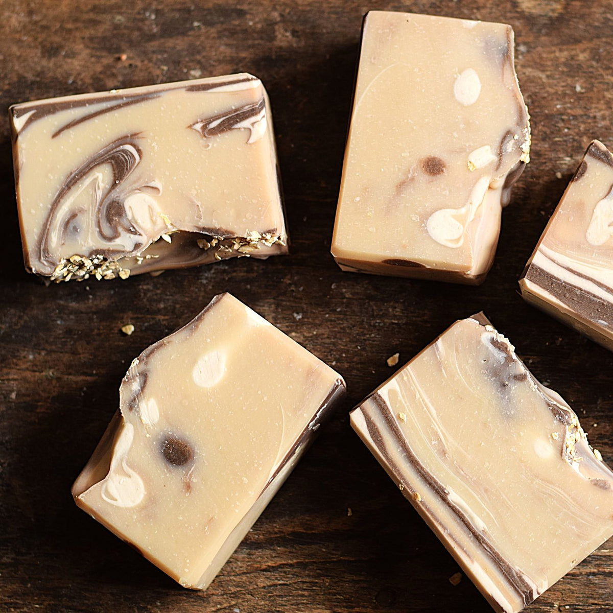 Five rectangular bars of artisanal soap with a creamy beige base and dark brown swirls, arranged on a dark rustic wooden surface.