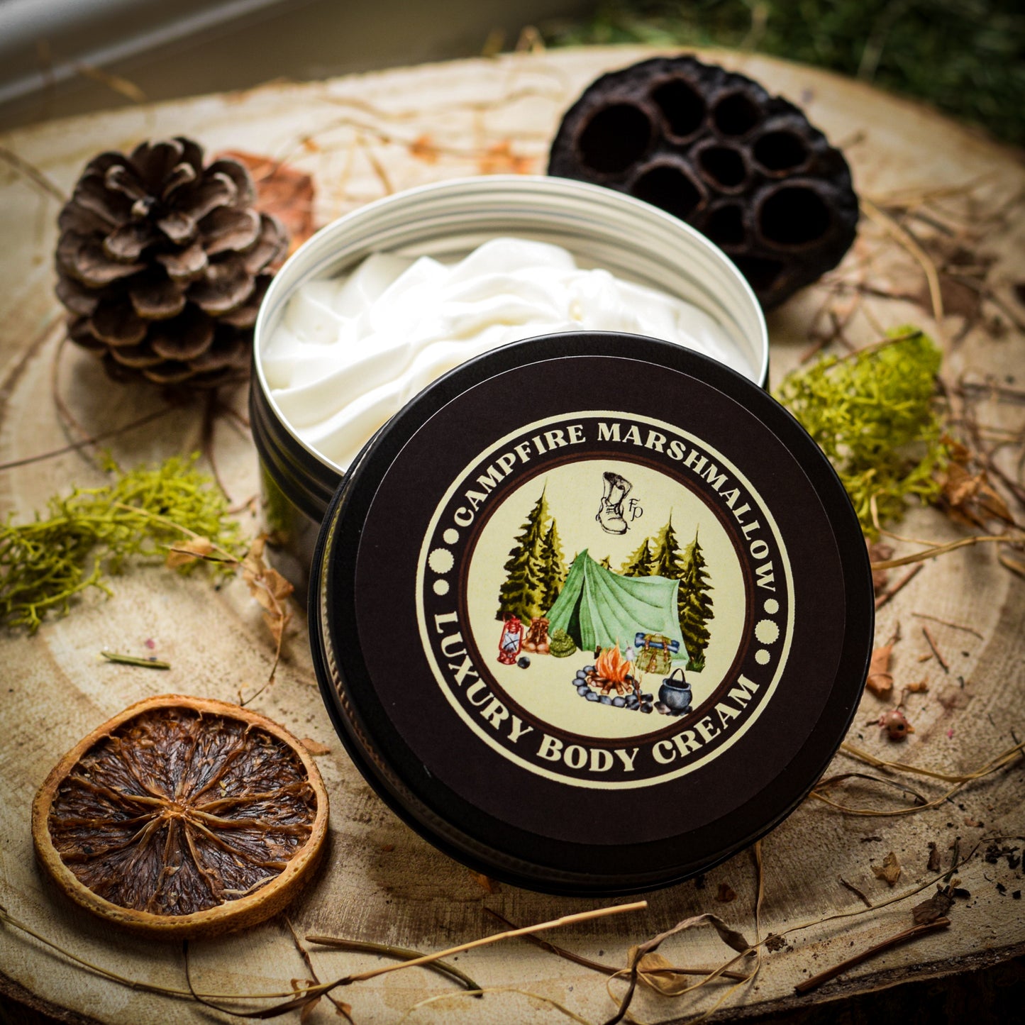 Open container of Campfire Marshmallow Luxury Body Cream on a rustic surface with natural elements.