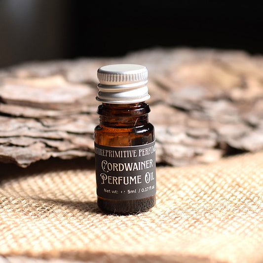 A small amber glass bottle of Cordwainer Perfume Oil by Futureprimitive soap, featuring a silver ribbed cap and a black label, set on a rustic burlap surface with a blurred wood background.