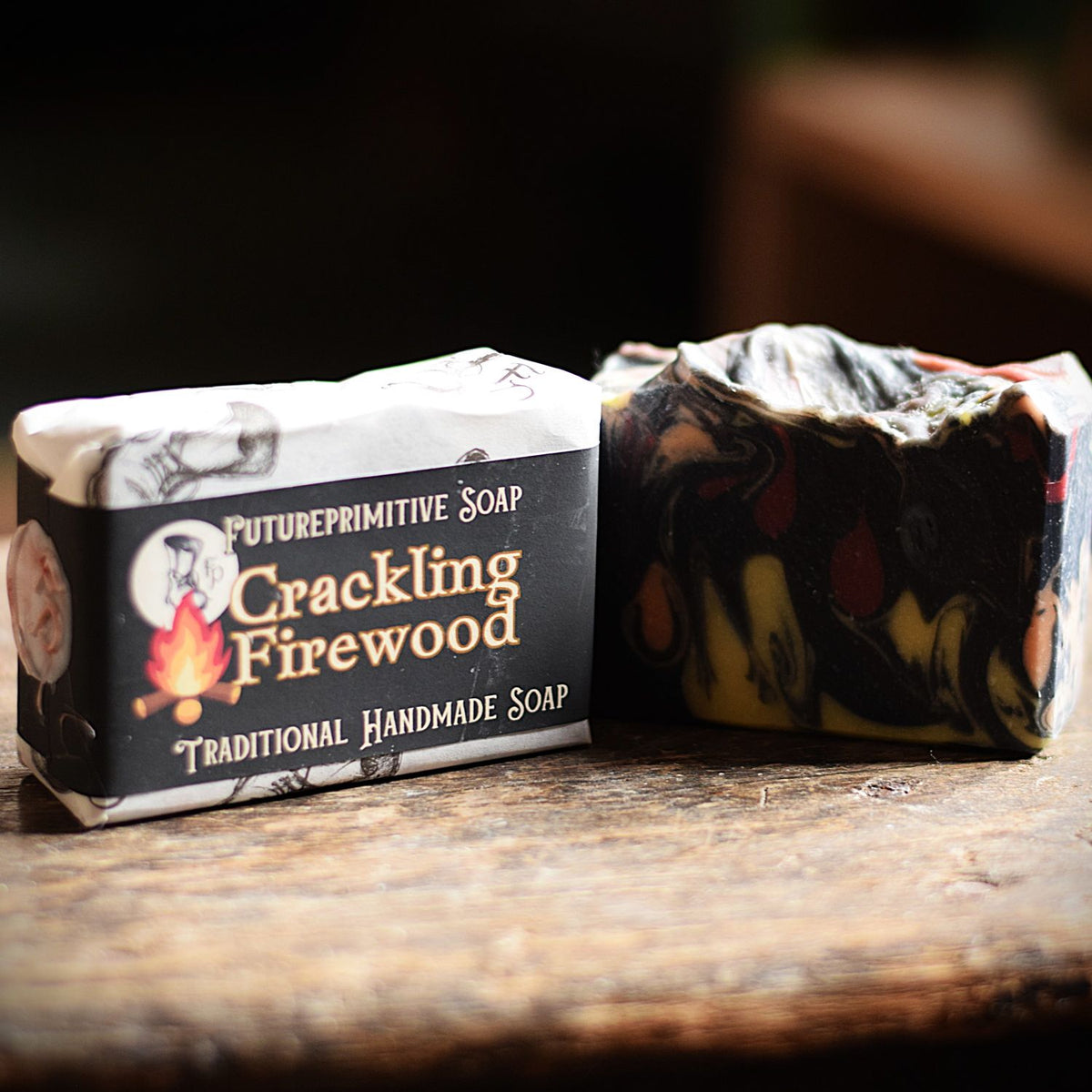 A bar of handmade soap in its paper wrapper sits next to an unwrapped marbled soap bar on a rustic wooden surface. The wrapper is black and white with the brand name 'FUTUREPRIMITIVE SOAP' and scent name 'Crackling Firewood'.