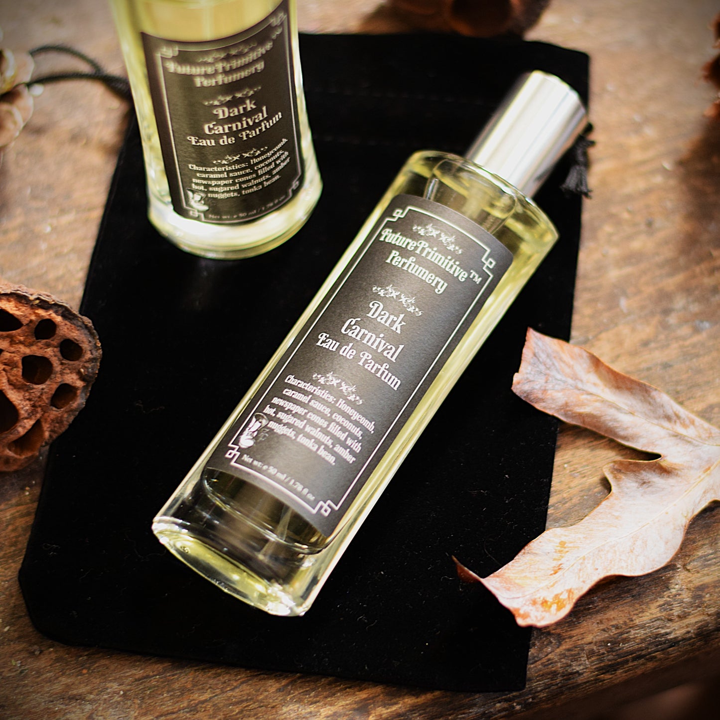 Two bottles of 'Dark Carnival Eau de Parfum' on a dark surface with dried leaves.