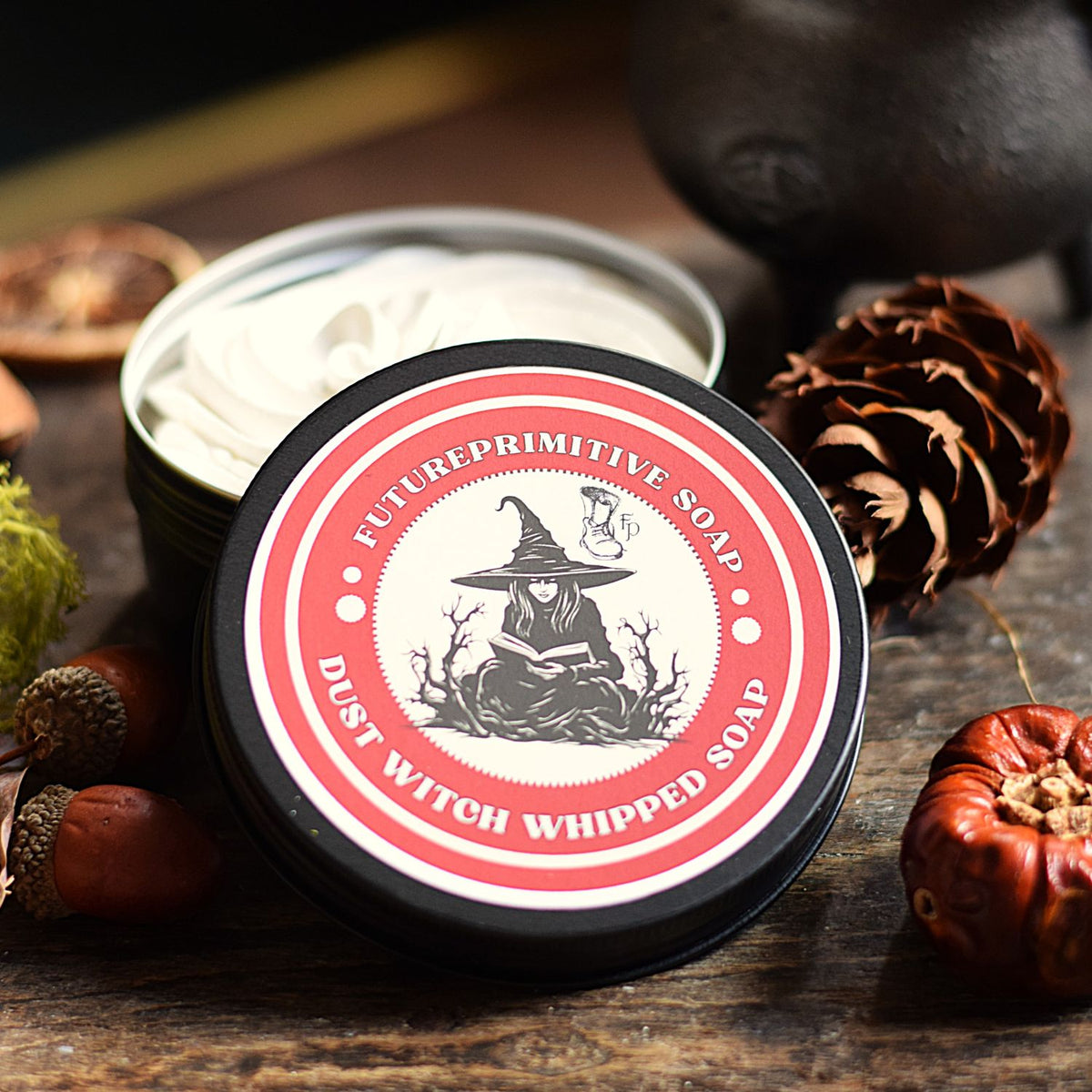 A round tin of Future Primitive Soap 'Dust Witch Whipped Soap' sits on a rustic wooden surface surrounded by autumn-themed props like acorns, a pinecone, and dried herbs. The lid features a black, white, and red design with an illustration of a witch reading.