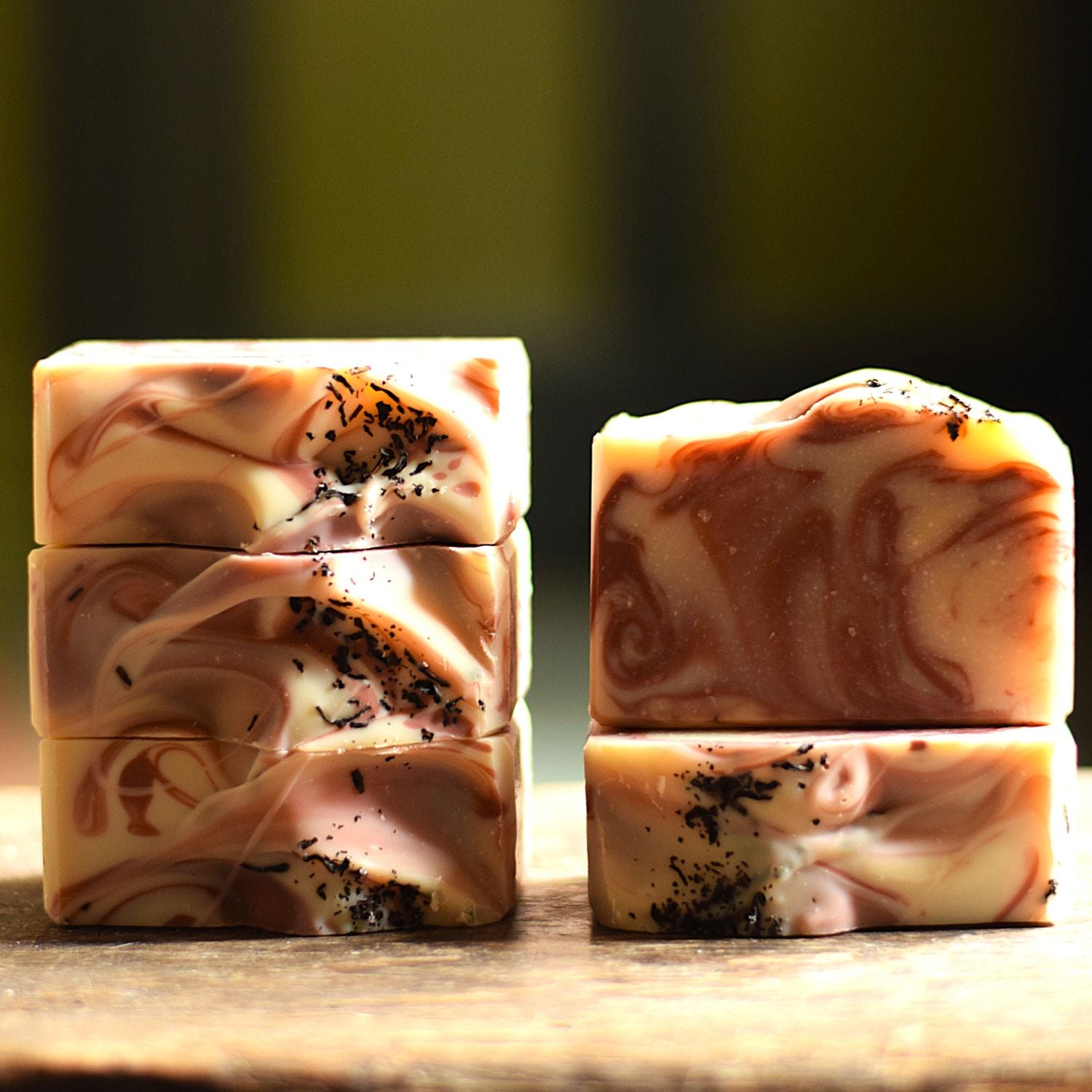 Four rectangular artisan soap bars with marbled cream, pink, and brown swirls, some topped with dark speckled flakes, displayed on a wooden surface against a blurred dark olive background.