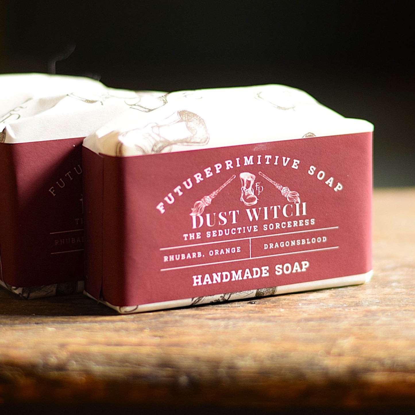 A handmade soap bar wrapped in a deep burgundy paper sleeve with white text reading 'Dust Witch' and 'Future Primitive Soap,' photographed on a rustic wooden surface with a dark blurred background.