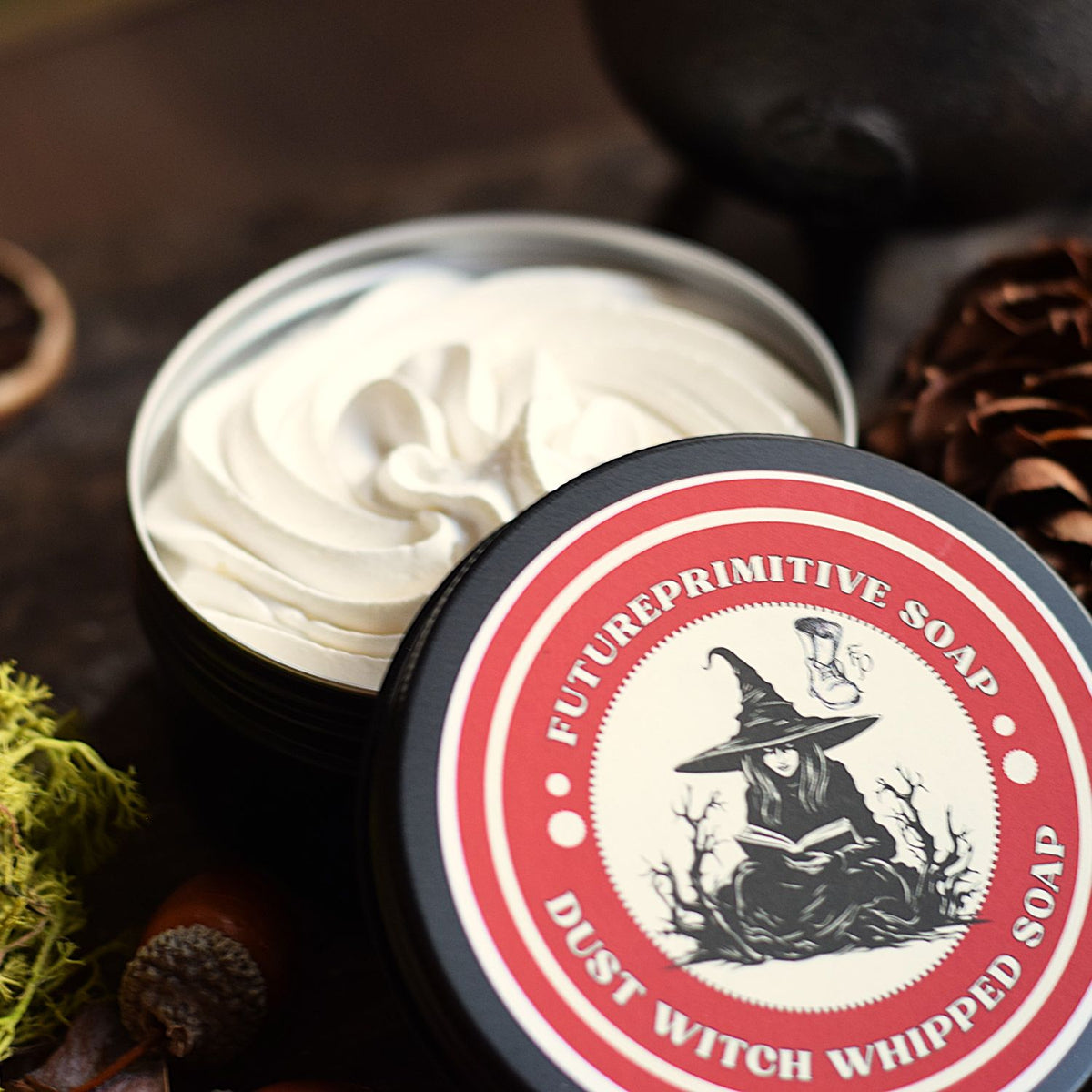 A tin of whipped soap with the lid off, showing the creamy off-white product inside. The lid features a black, red, and white label with a witch illustration and the text 'FUTUREPRIMITIVE SOAP' and 'DUST WITCH WHIPPED SOAP'.