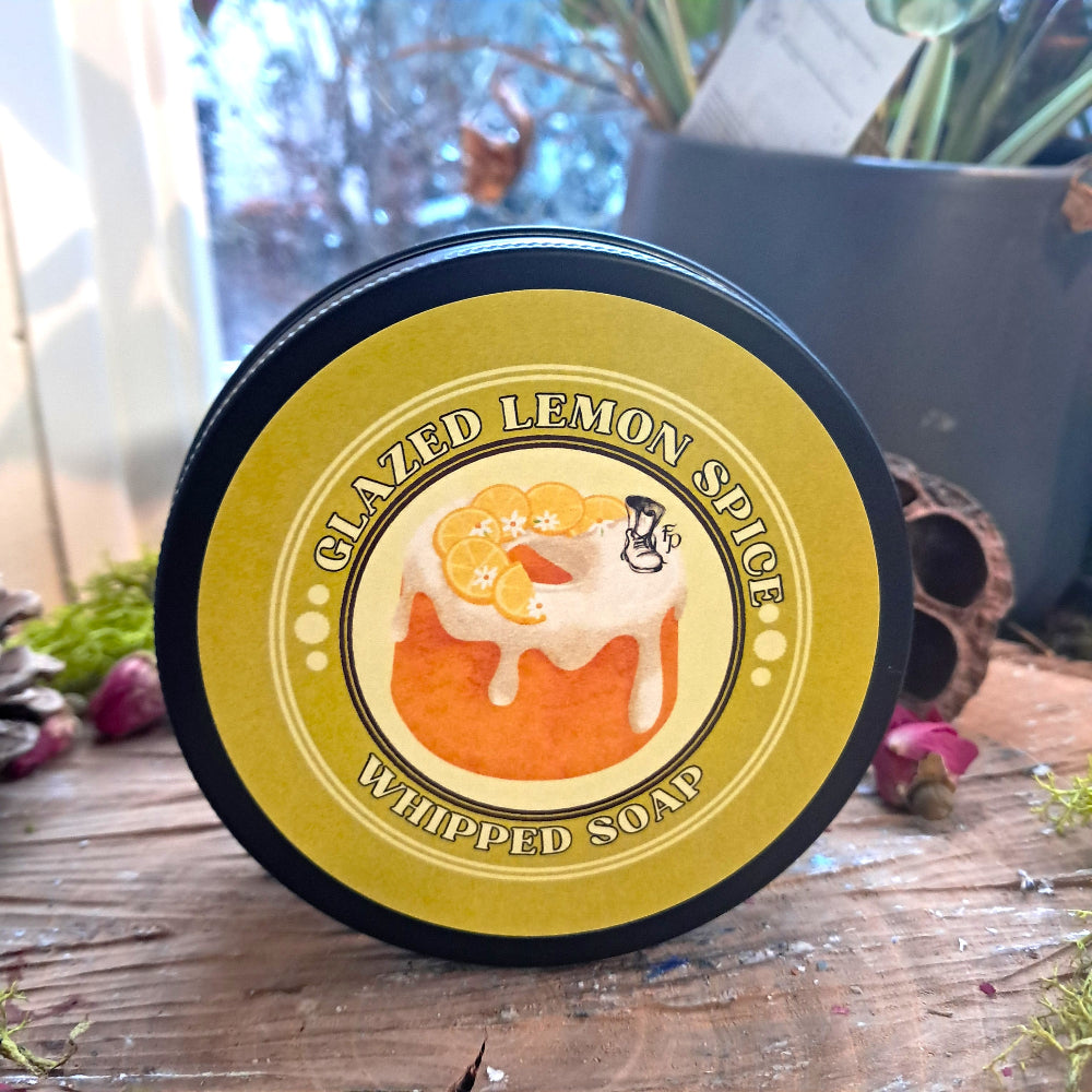 Container of glazed lemon spice whipped soap on a wooden surface with plants in the background
