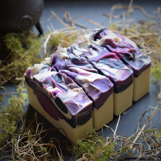 Four rectangular bars of artisanal soap with a marbled swirl pattern of purple, cream, and black, arranged on a dark slate surface with dried grass and moss.