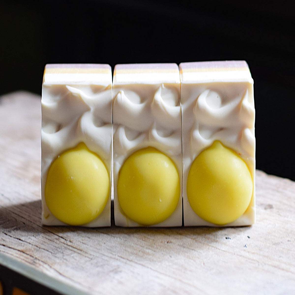 lemon handmade soap with lemon soaps on the top of each bar. the soap is sitting on a rustic wooden surface with a black background.