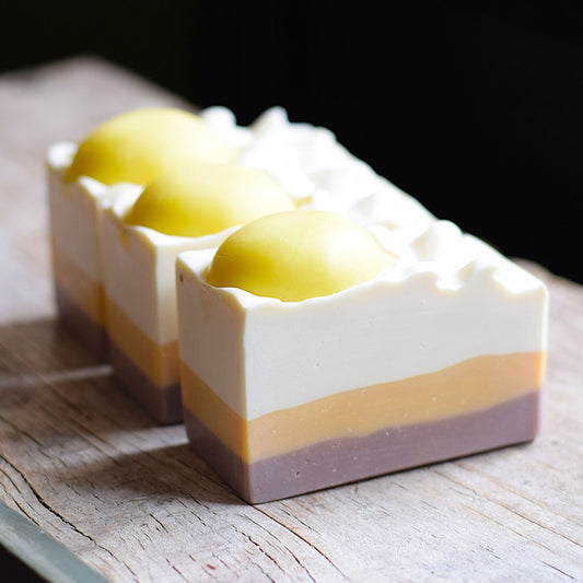 a soap that looks like lemon meringue pie. it is sitting on a wooden surface with a black blurred background.