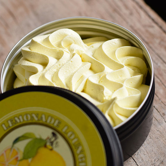 A close-up of a whipped cream soap swirled in an open round metal tin, with the branded lid placed in front on a rustic wooden surface. The label reads 'LEMONADE LOUNGER' and shows lemon imagery.