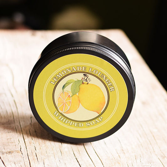 A round tin of Lemonade Lounger Whipped Soap, featuring a black metal container and a yellow label with illustrations of lemons, set on a light wooden surface.