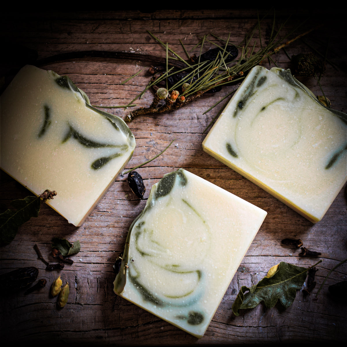 Three rectangular bars of handmade soap with green leaf swirls on a textured dark wood background, surrounded by dried botanicals.