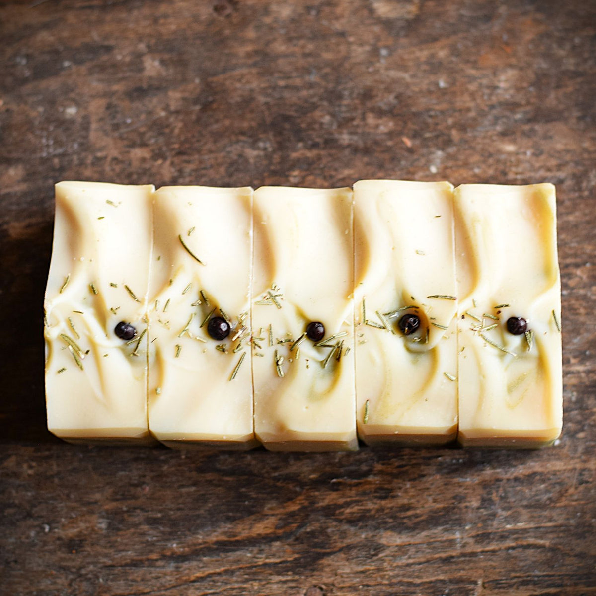 Five rectangular bars of handmade soap arranged side by side on a rustic dark wooden surface. The soap is a warm creamy beige color with swirls of olive oil and small dark botanical inclusions.